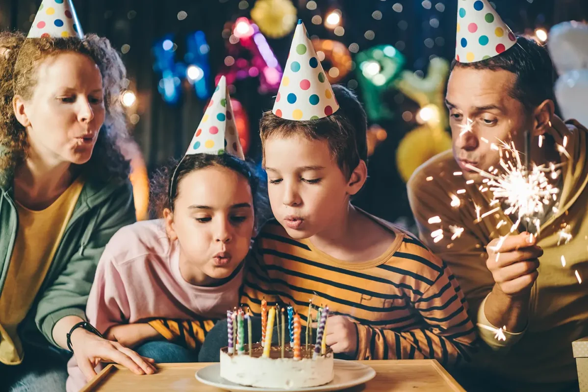 Familie purtând pălării de petrecere suflă în lumânările de pe tort și țin scânteietoare la o petrecere aniversară îmbelșugată.